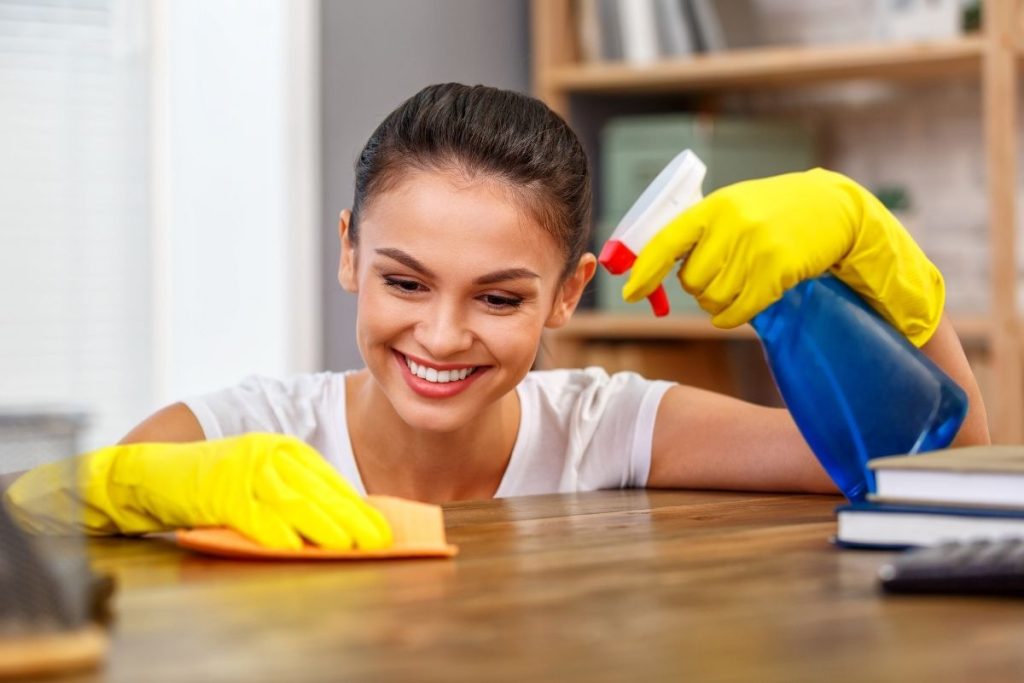 A woman in yellow rubber gloves cleans a desk, focused on removing dust and organizing items.