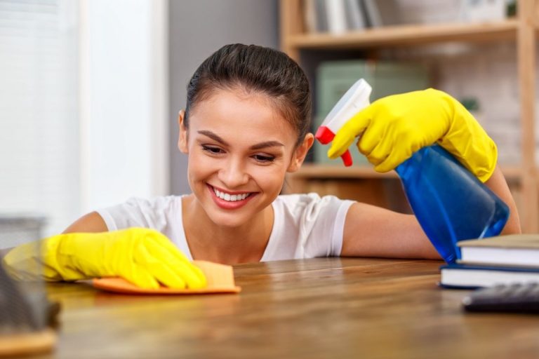 A woman in yellow rubber gloves cleans a desk, focused on removing dust and organizing items.